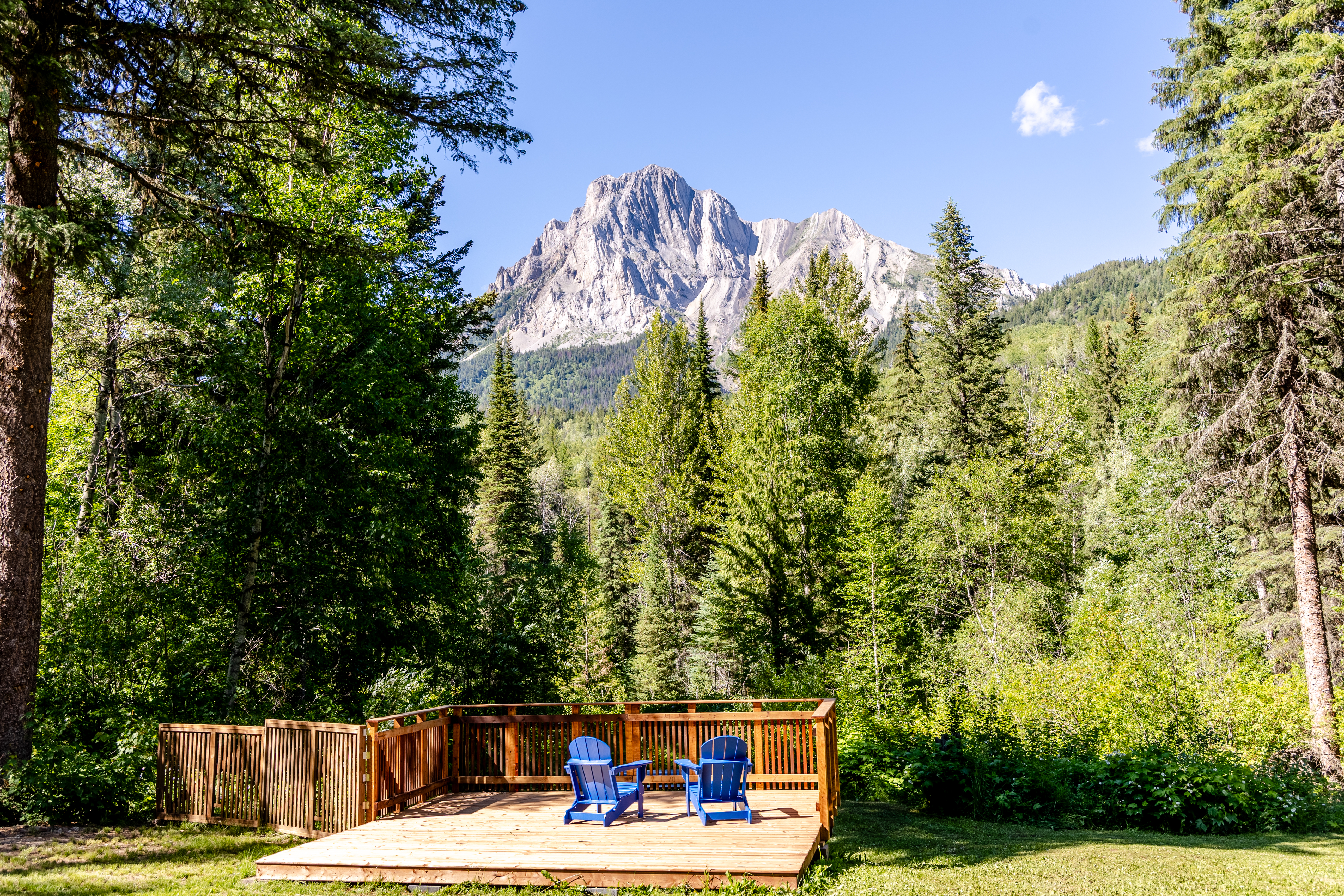 A wooden deck with two chairs is situated in a backyard, surrounded by trees and a mountain in the background.